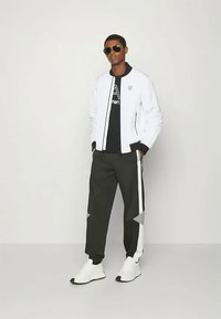 Giacca bomber bianca sopra una t-shirt nera, abbinata a pantaloni della tuta scuri con strisce laterali bianche e accenti grigio chiaro, indossata con occhiali da sole.