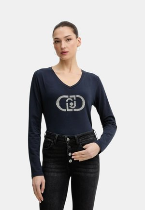 Donna con capelli scuri raccolti in uno chignon, che indossa una maglia blu scuro a maniche lunghe con scollo a V e logo di strass, jeans neri a vita alta, in piedi contro sfondo bianco.