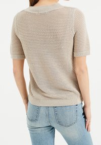 Beige stickad tröja med korta ärmar, med horisontell ribbad struktur, rund halsringning och en figursydd midja. Bärs med ljusblå jeans.