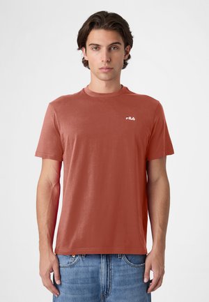 Jonge man met kort bruin haar, gekleed in een roestkleurig FILA T-shirt en blauwe jeans, staand tegen een effen witte achtergrond.