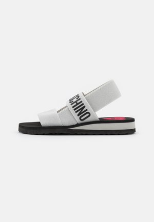 Hvid sandal med sort sål, elastiske remme inklusive en med teksten "MOSCHINO", og en lyserød logo på indersålen.