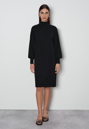 Schwarzes Strickkleid mit hohem, gewelltem Ausschnitt, langen Ballonärmeln und lockerer Passform. Getragen mit schwarzen, strapschuhen.