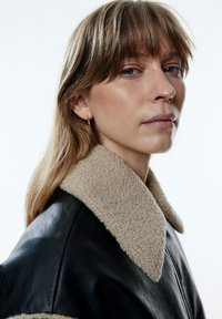 Schwarze Lederjacke mit einem beigen Schaffellkragen; verfügt über ein elegantes Design, eine glatte Textur und einen Reißverschluss. Nahaufnahme des Kragens.