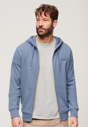 Felpa con zip blu realizzata in tessuto morbido, con cappuccio regolabile e tasca frontale, presenta un logo ricamato sul petto. Indossata sopra una maglietta grigia.