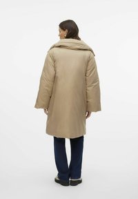 Beige pufferjakke med høj krave, oversized pasform og lange ærmer, der har en glat overflade og en knælang længde. Bæres med mørke jeans.