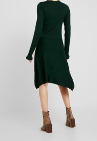 Grön långärmad klänning med ojämn nederkant och ribbad textur, kombinerad med bruna klackstövlar med ormskinnsmönster.