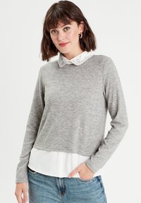 Graues Langarmoberteil mit einem weißen Kragenhemd darunter. Der Kragen hat zarte Stickereien. Lässiger Schnitt, weiche Textur und geschichtetes Design.