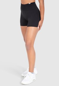 Shorts neri a vita alta, senza cuciture, texture liscia, design aderente, che raggiungono la metà della coscia. Abbinati a sneaker bianche per contrasto.