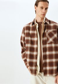 Camicia overshirt a quadretti marrone e crema realizzata in tessuto morbido, con chiusura frontale a bottoni, tasca sul petto e vestibilità rilassata con maniche lunghe.