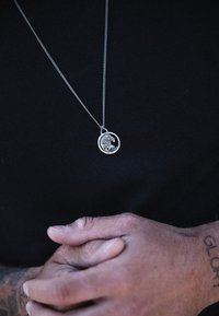 Silverhänge med vågdesign på en tunn kedja, bärs över en svart tröja. Händer sammanlänkade nedanför, med texturerad hud och tatueringar synliga.