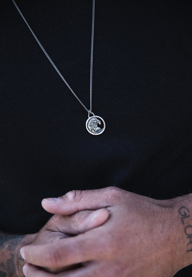 Silverhänge med vågdesign på en tunn kedja, bärs över en svart tröja. Händer sammanlänkade nedanför, med texturerad hud och tatueringar synliga.