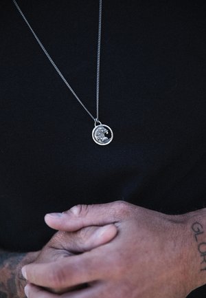Silver chain necklace with a round pendant featuring a wave design; worn on a dark shirt, hands clasped, showcasing textured metal.