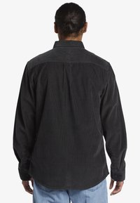 Camisa de pana negra con mangas largas, cuello abotonado y yugo trasero. Presenta una textura de canalé vertical y puños con botón.