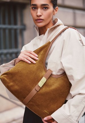 Bolso hobo de ante marrón con una correa de cuero beige y un cierre dorado. Presenta una textura suave y una forma redondeada y espaciosa.