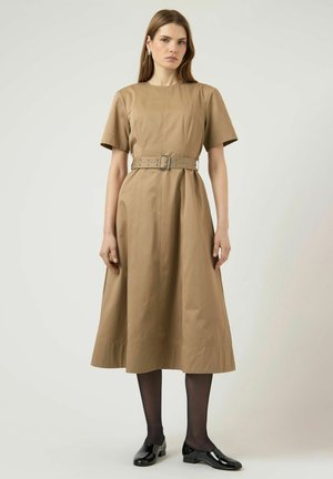 Vrouw staat in een zandkleurige midi-jurk met ceintuur en korte mouwen, zwarte doorschijnende panty's en zwarte lakleren loafers tegen een effen achtergrond.