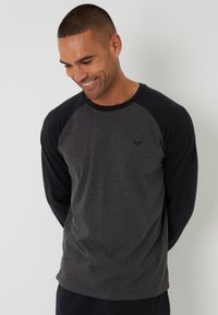 Camisa de raglán de manga larga con cuerpo gris y mangas negras, hecha de una tela suave, con un pequeño logo en el pecho. Textura suave.