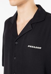 Chemise noire à manches courtes avec col, dotée d'une fermeture à boutons sur le devant et d'un logo blanc "PEGADOR" sur le côté gauche. Texture de tissu lisse.