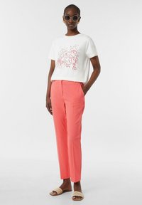 Weißes Baumwoll-T-Shirt mit Hummer-Grafik, kombiniert mit korallfarbenen, maßgeschneiderten Hosen. Das Modell trägt übergroße, runde Sonnenbrillen und beigefarbene Sandalen.