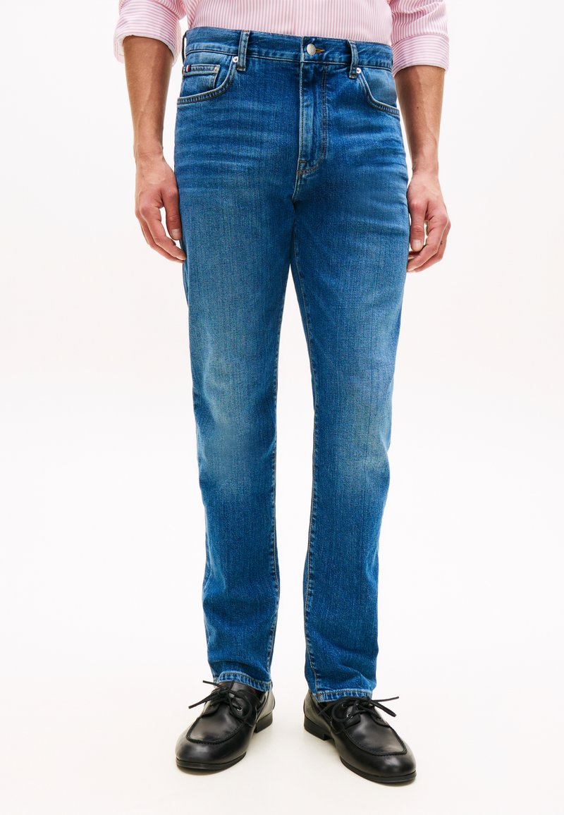 Άνδρας που φοράει μπλε τζιν με ίσια μπατζάκια, μαύρα δερμάτινα παπούτσια και ανοιχτό ροζ ριγέ πουκάμισο με τα μανίκια σηκωμένα.