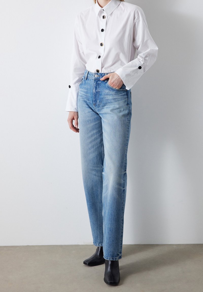 Jean en denim bleu clair à coupe droite, associé à une chemise blanche à boutons noirs et manches longues. Bottines noires.