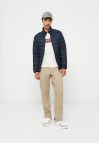 Mørkeblå puffjakke, cremefarvet t-shirt med logo, beige bukser og hvide sneakers. Modellen bærer en lysegrå kasket. Enkel design, funktionel stil.
