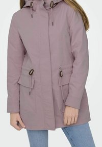 Kobieta w fioletowej kurtce z kapturem i sznurkami, z klapkami w kieszeniach, w połączeniu z jasnoniebieskimi dżinsami na białym tle.