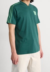 Green cotton t-shirt with a v-neck, featuring green three-stripe design on sleeves and a small logo on the chest. Smooth texture.