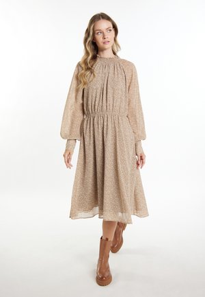 Femme aux longs cheveux ondulés portant une robe midi marron clair à motifs avec manches longues et des bottines marron, debout sur un fond blanc.