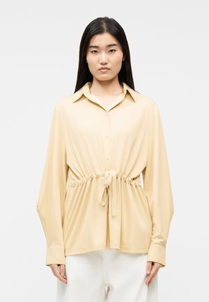 Γυναίκα που φοράει μπεζ πουκάμισο με κουμπιά, με κορδόνι στη μέση, μακριά μανίκια και λευκό παντελόνι, στέκεται μπροστά σε μια απλή ανοιχτόχρωμη επιφάνεια.