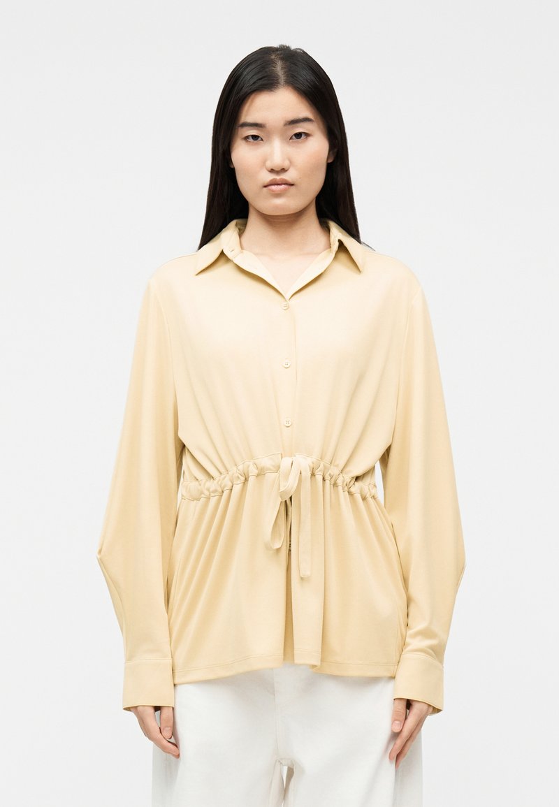 Γυναίκα που φοράει μπεζ πουκάμισο με κουμπιά, με κορδόνι στη μέση, μακριά μανίκια και λευκό παντελόνι, στέκεται μπροστά σε μια απλή ανοιχτόχρωμη επιφάνεια.