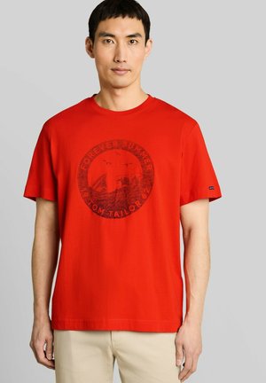 Mann trägt ein rotes T-Shirt mit kurzen Ärmeln, rundem Grafik- und Textmotiv, kombiniert mit hellbeigen Hosen, steht vor einem schlichten Hintergrund.