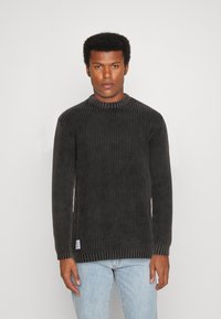 Schwarzer, gerippt gestrickter Pullover mit rundem Halsausschnitt und langen Ärmeln, versehen mit einem strukturierten Stoff und einem kleinen Markenetikett am Saum.