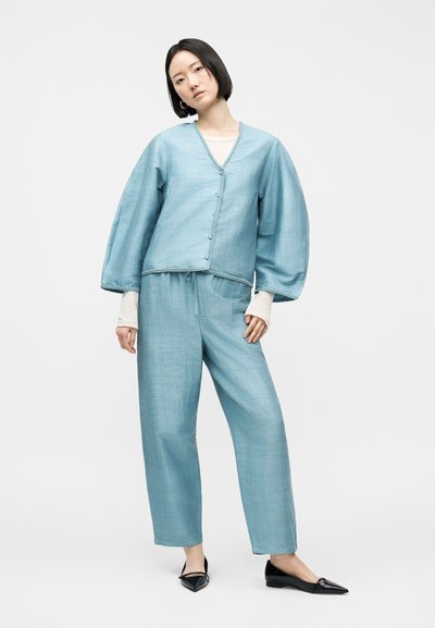 Γυναίκα που φοράει ανοιχτό μπλε υφασμάτινο σακάκι με φαρδιά μανίκια και αντίστοιχο φαρδύ παντελόνι, συνδυασμένα με μαύρα μυτερά παπούτσια σε λευκό φόντο.