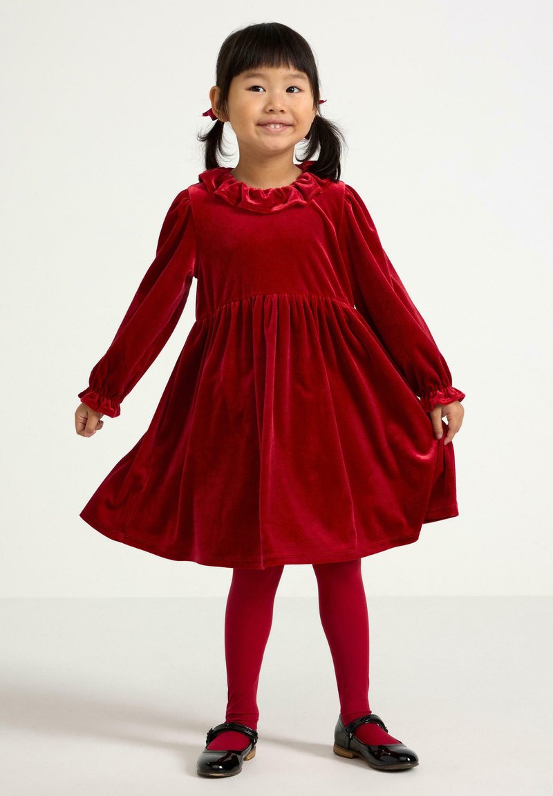 Czerwona aksamitna sukienka z falbaniastym dekoltem, długimi rękawami i rozkloszowanym dołem. Uzupełniona czerwoną rajstopami i czarnymi lakierowanymi butami.