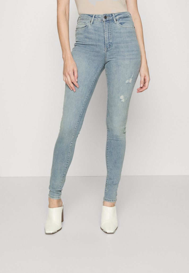 Femme portant des jeans skinny déchirés bleu clair et des chaussures à talons blanches à enfiler, se tenant devant un fond simple clair.