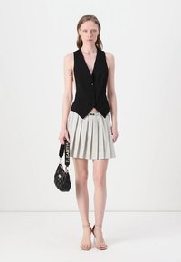 Gilet nero senza maniche con bottoni frontali, abbinato a una gonna plissettata con pois bianchi. Tiene in mano una borsa nera con texture.