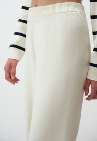 White, loose-fitting lounge pants made of a soft fabric, featuring a ribbed waistband and a smooth texture, shown from the side.