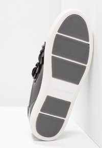Sneaker en cuir noir avec surface texturée, doté d'une semelle en caoutchouc blanc et d'un grip à motif distinct, affichant un accent en nœud sur le côté.