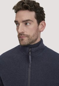 Man met kort, krullend bruin haar en een getrimd baardje, gekleed in een marineblauwe zip-up trui, kijkt naar links tegen een effen achtergrond.
