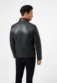 Schwarze Lederjacke mit hohem Kragen und schmalem Schnitt. Merkmale sind sichtbare Nähte und ein Schnallen-Detail an der Taille. Glatte Textur.