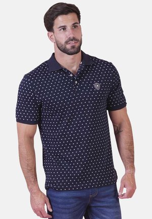 Homme portant un polo bleu marine avec de petits motifs en forme de losange blanc et un jean bleu, debout devant un fond uni.