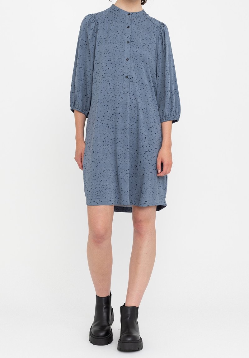 Abito blu, lungo fino al ginocchio, con colletto tondo, maniche a sbuffo e motivo a pois neri. Presenta una chiusura a bottoni sul davanti. Indossato con stivali neri alla caviglia.