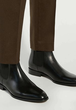 Black leather Chelsea boots with a smooth finish, elastic side panels, pointed toe, and flat sole, paired with brown tailored pants.