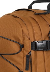 Ruskea kangasreppu, jossa on mustia yksityiskohtia, säädettävät hihnat, vetoketjulliset taskut ja muovinen solki turvallista sulkemista varten.