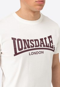 Cremefarvet bomulds t-shirt med et stort maroon "LONSDALE" logo og teksten "LONDON" nedenunder i en fed, moderne skrifttype.