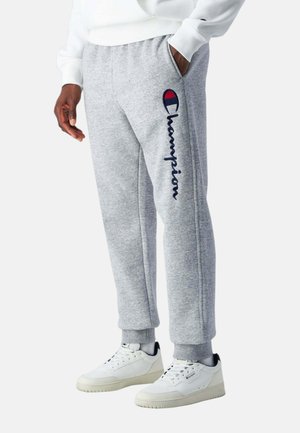 Persona che indossa pantaloni della tuta Champion grigio chiaro con logo sulla gamba e scarpe da ginnastica bianche, in piedi con una mano in tasca.