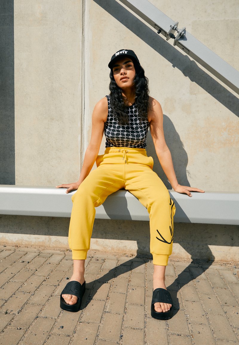 Melnbalts raksts bez piedurknēm, dzelteni sporta bikses ar sānu dizainu un melni slaidi sandalītes. Modelis sēž uz soliņa pret sienu.