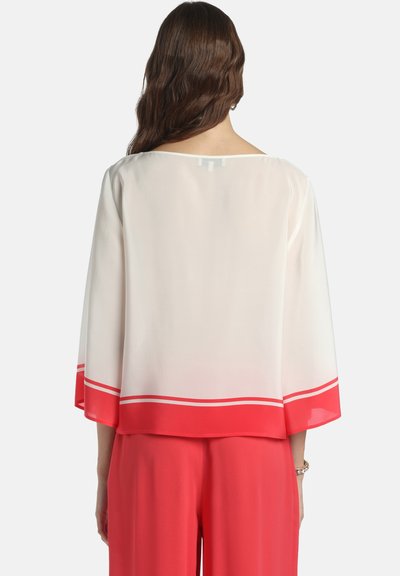 Femme aux longs cheveux bruns portant un chemisier blanc ample avec des bordures rouges et un pantalon large assorti rouge, debout de dos.