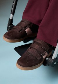 Καφέ σουέτ παπούτσια με αυτοκόλλητες ταινίες, μπεζ λεπτομέρειες και σόλα από καουτσούκ, τοποθετημένα σε υποπόδιο αναπηρικής καρέκλας με μπορντό παντελόνι.