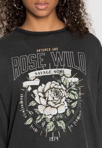 Black long-sleeve shirt with a white and green rose graphic, gold and white text, round neckline, and relaxed fit.